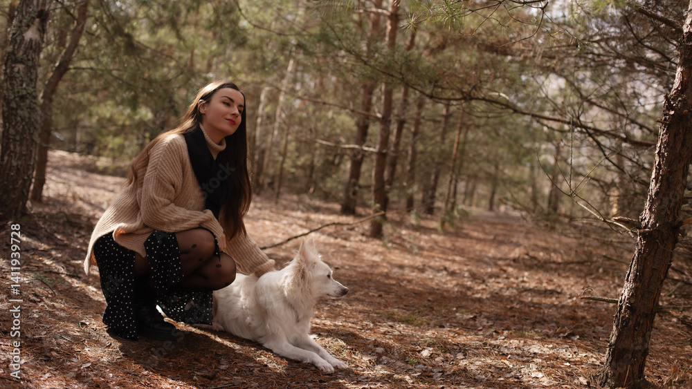 A woman in a cozy sweater and black tights with polka dots kneels beside her white dog in a forest. They share a peaceful moment, surrounded by nature and soft sunlight filtering through the trees.