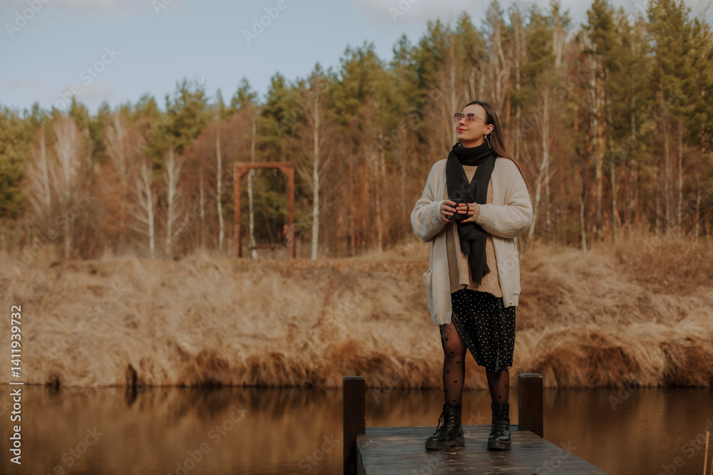 A woman standing on a wooden dock by a serene pond, holding a cup and looking up, enjoying the peaceful surroundings. She wears a cozy sweater, black tights with polka dots, and stylish boots.