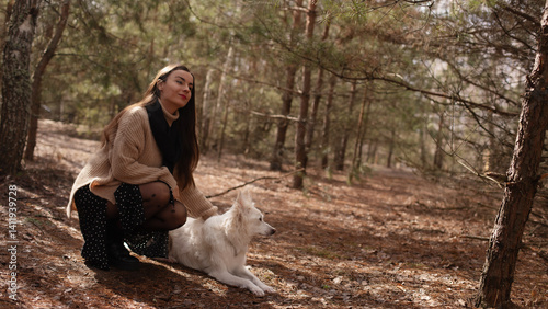 Obraz na płótnie A woman in a cozy sweater and black tights with polka dots kneels beside her white dog in a forest. They share a peaceful moment, surrounded by nature and soft sunlight filtering through the trees. w salonie