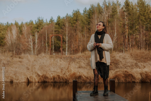 Obraz na płótnie A woman standing on a wooden dock by a serene pond, holding a cup and looking up, enjoying the peaceful surroundings. She wears a cozy sweater, black tights with polka dots, and stylish boots. w salonie