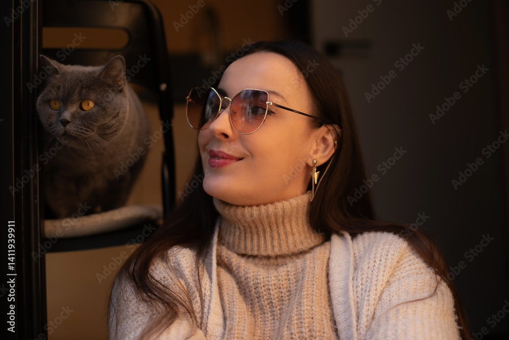 A woman in a cozy beige turtleneck sweater smiles softly, wearing stylish sunglasses. A grey cat sits beside her on a chair, creating a warm and peaceful indoor moment with a soft, intimate atmosphere
