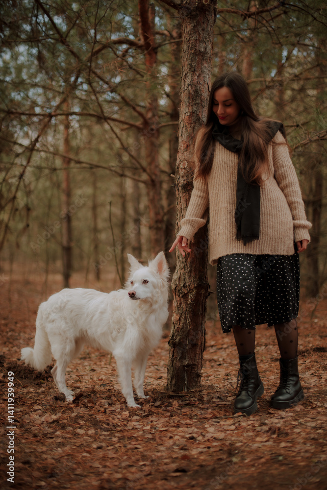 A woman in a cozy beige sweater and black skirt stands in the forest, smiling and gently interacting with her white dog. The scene is calm, with autumn leaves and a peaceful natural backdrop.