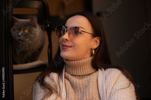 A woman in a cozy beige turtleneck sweater smiles softly, wearing stylish sunglasses. A grey cat sits beside her on a chair, creating a warm and peaceful indoor moment with a soft, intimate atmosphere