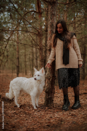 Obraz na płótnie A woman in a cozy beige sweater and black skirt stands in the forest, smiling and gently interacting with her white dog. The scene is calm, with autumn leaves and a peaceful natural backdrop. w salonie