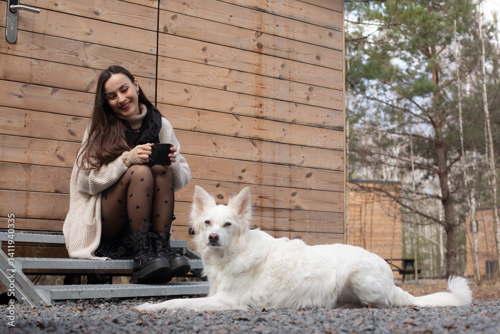 A woman sitting on steps outside a wooden cabin, holding a black cup and smiling, with her white dog lying beside her. The scene is peaceful, with natural surroundings and a relaxed atmosphere.