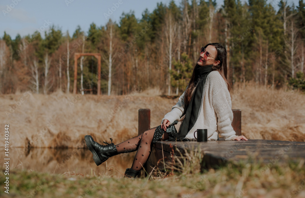 A woman sitting on a wooden dock by a calm pond, enjoying a coffee in the sun. She wears a cozy sweater, black tights with polka dots, and boots, smiling and surrounded by natural beauty.