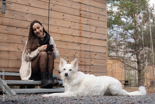 Obraz na płótnie A woman sitting on steps outside a wooden cabin, holding a black cup and smiling, with her white dog lying beside her. The scene is peaceful, with natural surroundings and a relaxed atmosphere. w salonie