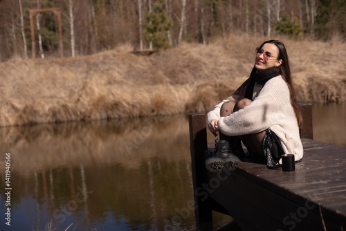 Obraz na płótnie A young woman sitting on a wooden dock by a peaceful pond, wearing a cozy sweater, black tights, and boots, enjoying a warm drink, smiling in the sunlight with a serene natural backdrop. w salonie