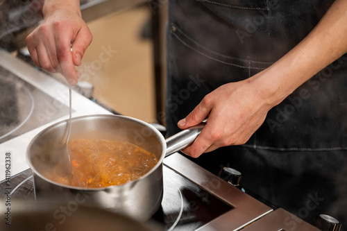 A close-up of a chef's hand stirring a simmering pot of stew on the stovetop, with steam rising, showcasing the cooking process in a professional kitchen.