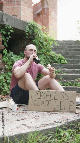 A homeless man with a sign asking for help eats a sandwich and drinks water. Vertical video.