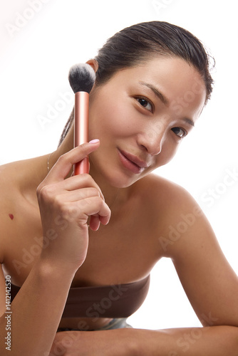 Obraz na płótnie Beautiful Korean woman in her 20s with clean, smooth skin holding makeup brush next to face on white background. Concept of cosmetic routine, natural beauty, wellness, makeup accessories w salonie