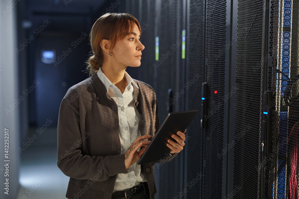 Fototapeta premium Asian woman with tablet checking server racks in data center illuminating from server lights. Modern technology environment with significant attention to equipment and cooling systems