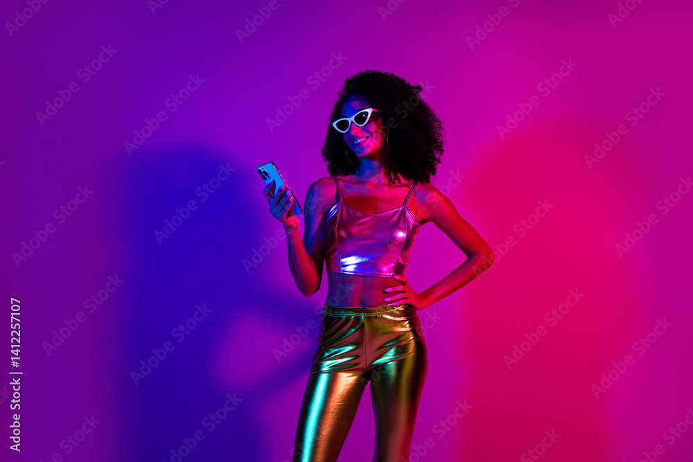 Trendy young woman in colorful neon lighting holding a phone and smiling confidently in a fashionable outfit Trendy young woman in colorful neon lighting holding a phone and smiling confidently in a fashionable outfit