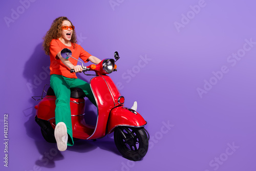Energetic young woman riding a bright red scooter on a vibrant purple background, showcasing a cheerful and fashionable lifestyle.