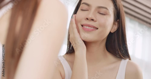Beautiful young woman applies moisturizer to her face while standing in front of a mirror.