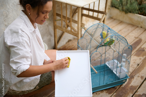 A woman carefully cleans the parts of a bird cage on an outdoor wooden terrace while her colorful birds remain perched inside, reflecting care and routine in pet maintenance.