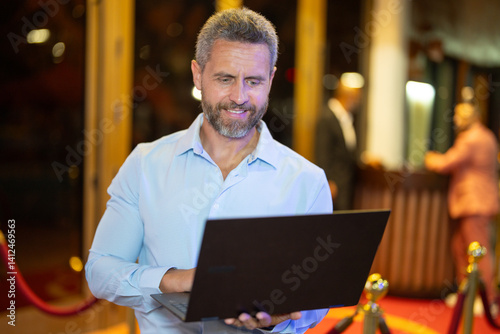 Business man using a laptop on night light. Business man work on laptop on street. Middle-aged business man planning work near night club. Business in 40s. Freelancer browsing on laptop outdoors.