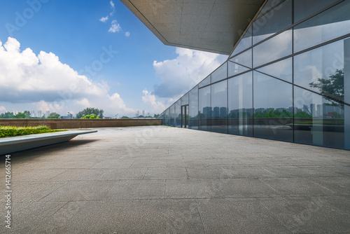 Fototapeta Naklejka Na Ścianę i Meble -  Empty stone floor on rooftop terrace beside modern glass wall building overlooking city skyline under blue sky background.
