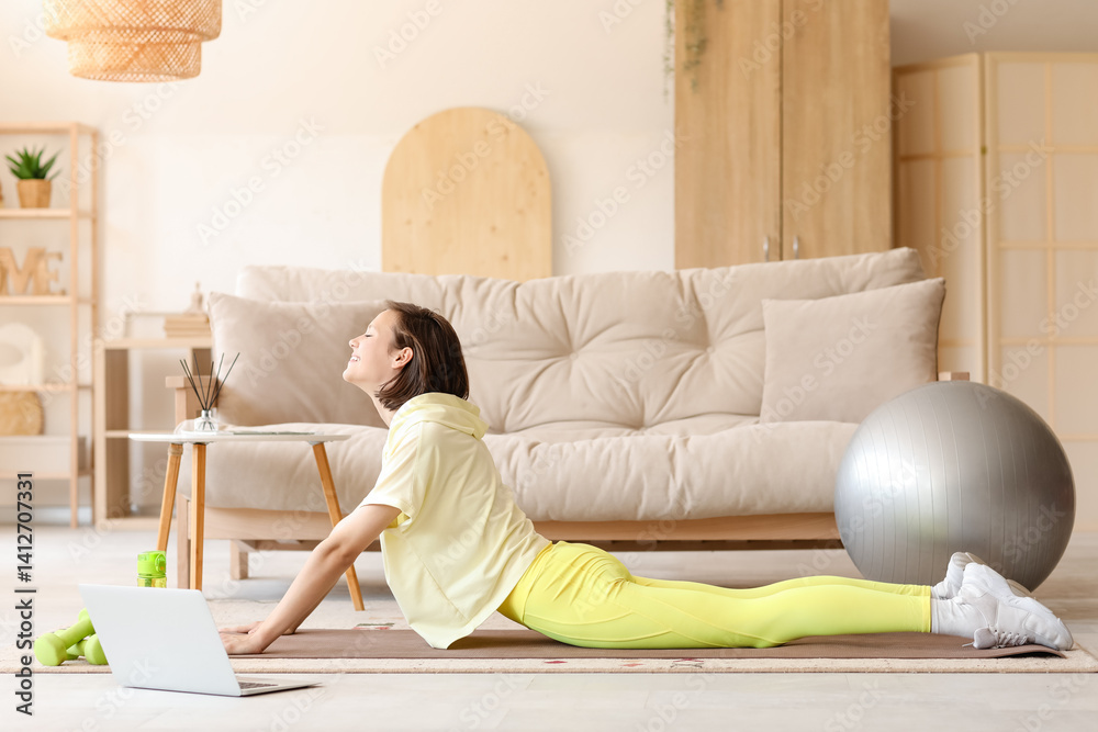 Beautiful young woman with laptop practicing yoga on mat in living room