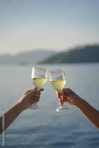 Two people clink wine glasses filled with white wine against serene lake backdrop, celebrating peaceful moment