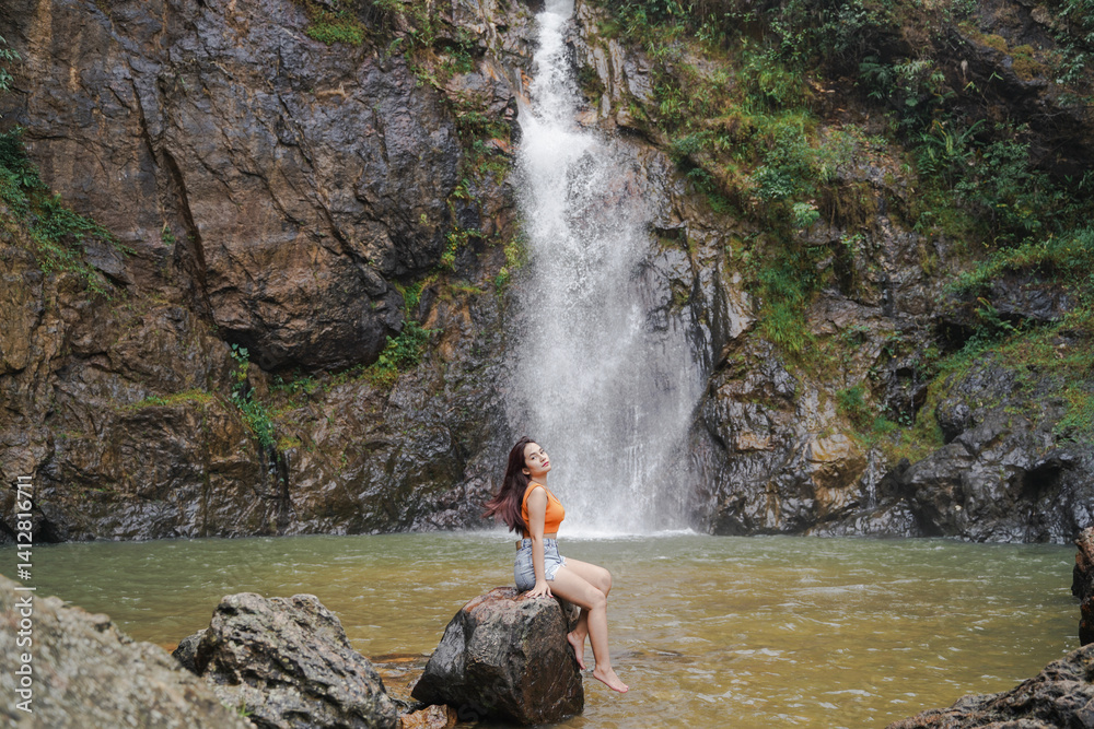 woman sits on rock by serene waterfall, surrounded by lush greenery and rocky cliffs