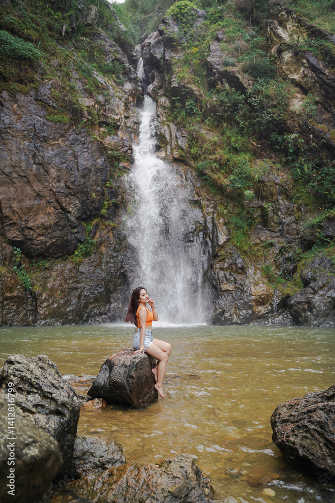 woman sits on rock by serene waterfall, surrounded by lush greenery and rocky cliffs