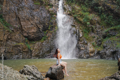 Obraz na płótnie woman sits on rock by serene waterfall, surrounded by lush greenery and rocky cliffs w salonie