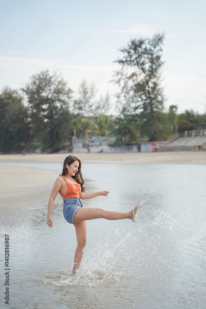 woman joyfully kicks water at beach, wearing bright orange top and denim shorts. serene background features trees and clear sky