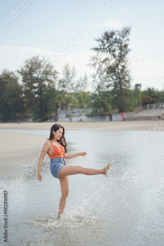 Obraz na płótnie woman joyfully kicks water at beach, wearing bright orange top and denim shorts. serene background features trees and clear sky w salonie