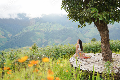 Obraz na płótnie woman sits peacefully on wooden platform under tree, overlooking lush green mountains and vibrant wildflowers w salonie
