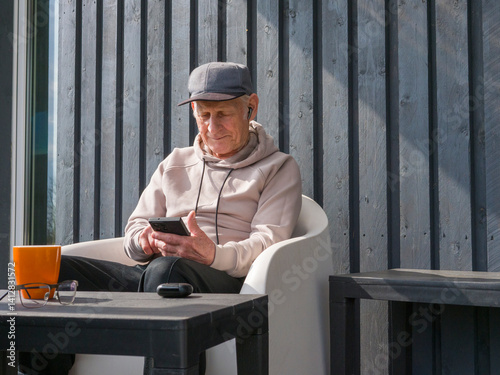 Sitting comfortably on a modern white chair on a wooden porch, a senior man wearing a baseball cap, glasses, and a light brown hoodie enjoys music through his earbuds while using his smartphone