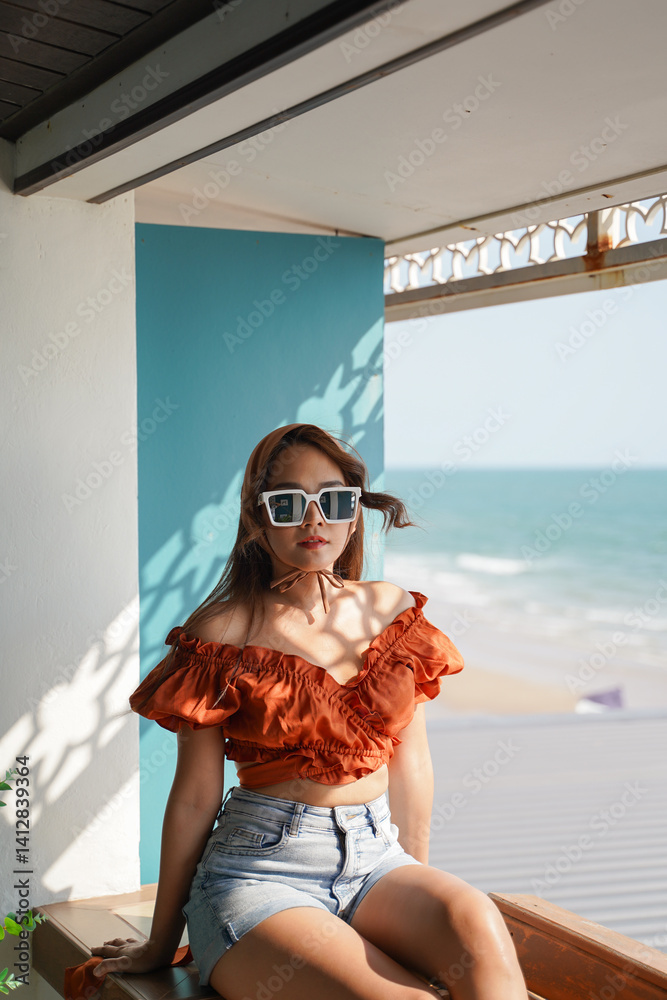 woman in stylish orange top and denim shorts enjoys sunny day by beach, wearing sunglasses