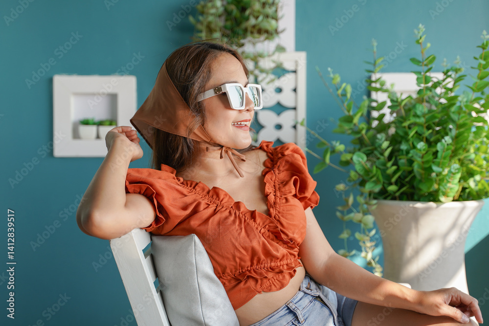 woman in stylish orange top and sunglasses enjoys sunny day, sitting on chair with blue wall and plants in background