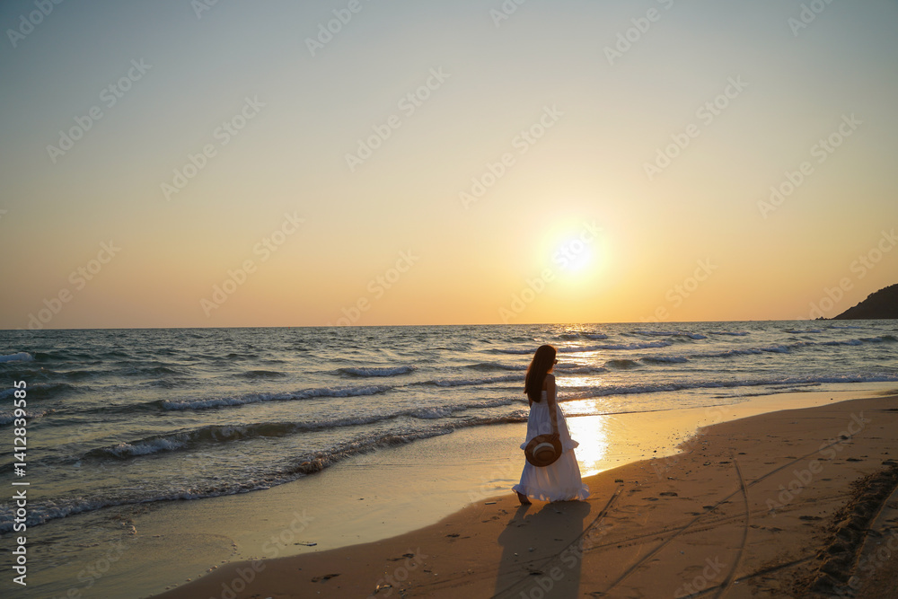 woman in white dress walks along serene beach at sunset, holding hat, with gentle waves lapping at shore