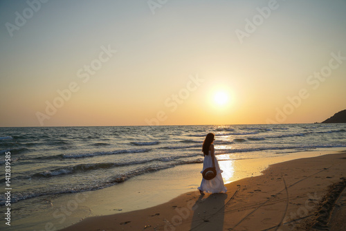 Obraz na płótnie woman in white dress walks along serene beach at sunset, holding hat, with gentle waves lapping at shore w salonie