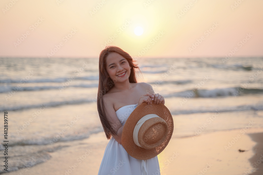 woman in white dress smiles joyfully while holding straw hat on beach at sunset