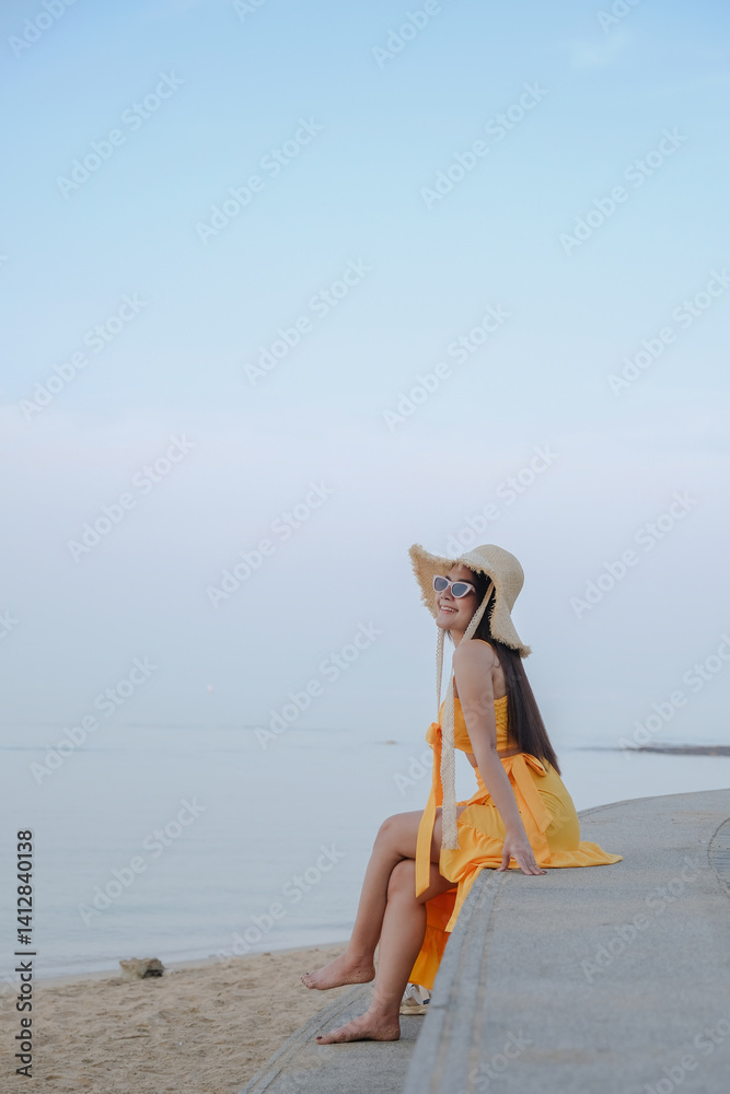 woman in yellow dress and sun hat sits on beach wall, enjoying serene ocean view