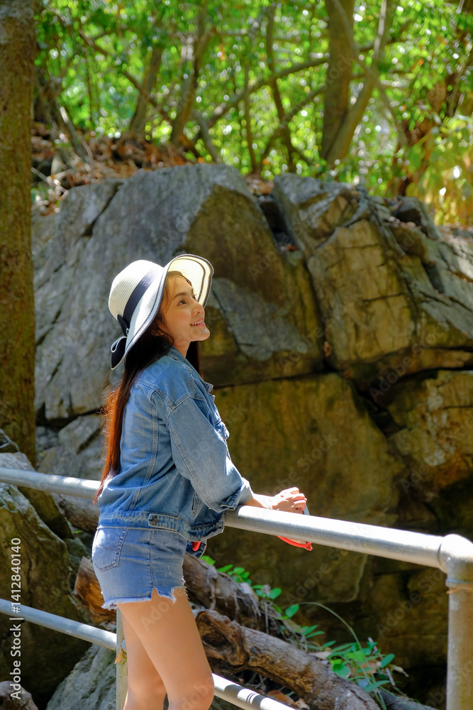 woman in denim outfit and sun hat enjoys sunny day in lush forest setting, leaning on railing with joyful expression