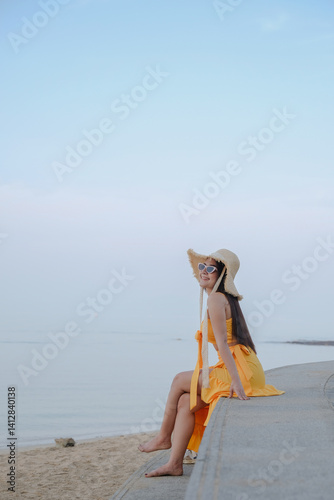 Obraz na płótnie woman in yellow dress and sun hat sits on beach wall, enjoying serene ocean view w salonie