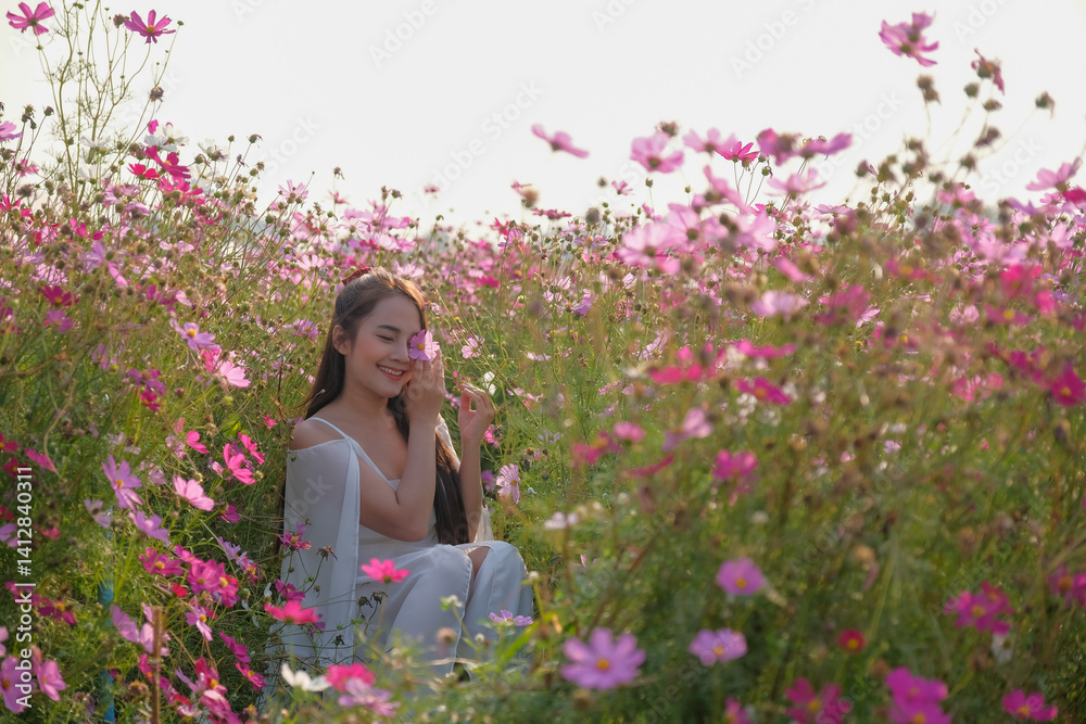 woman in white dress sits joyfully among vibrant pink and white flowers in sunlit field