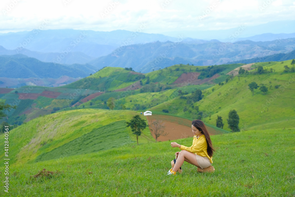 woman in yellow shirt sits on lush green hill, surrounded by rolling mountains and serene landscape