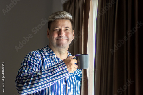 Smiling man with modern hairstyle in striped pajamas standing by the window with a mug