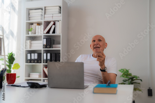 Senior businessman sitting at his desk in a white and bright office, having a video call on a laptop and explaining something raising a finger photo