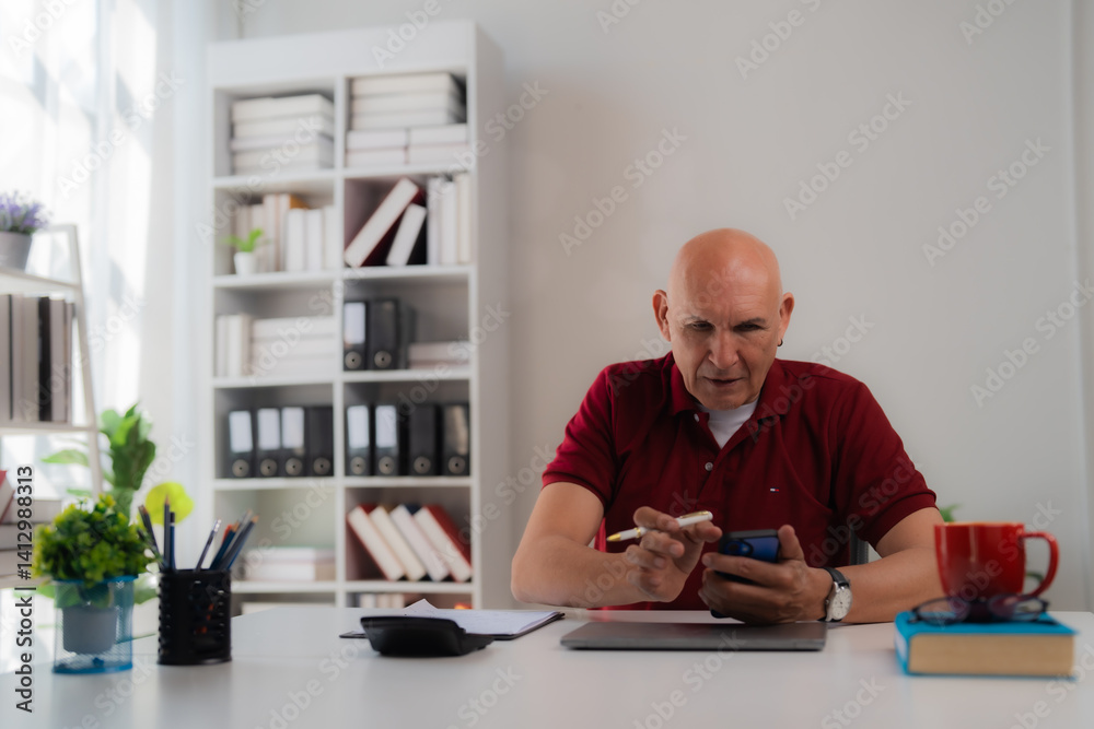 Senior manager sitting at desk and using smartphone with pen, working in bright modern office with bookshelves and plants, focused on his tasks Senior manager sitting at desk and using smartphone with pen, working in bright modern office with bookshelves and plants, focused on his tasks