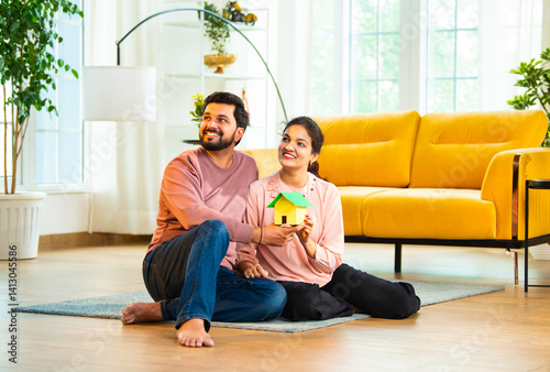 Smiling Asian couple holding colorful paper house model while planning new home in living room