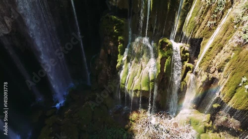 Sunlight filtering across moss-covered rocky terrain, powerful waterfall plunging through lush green turkish landscape, rushing beside steep mountain cliff