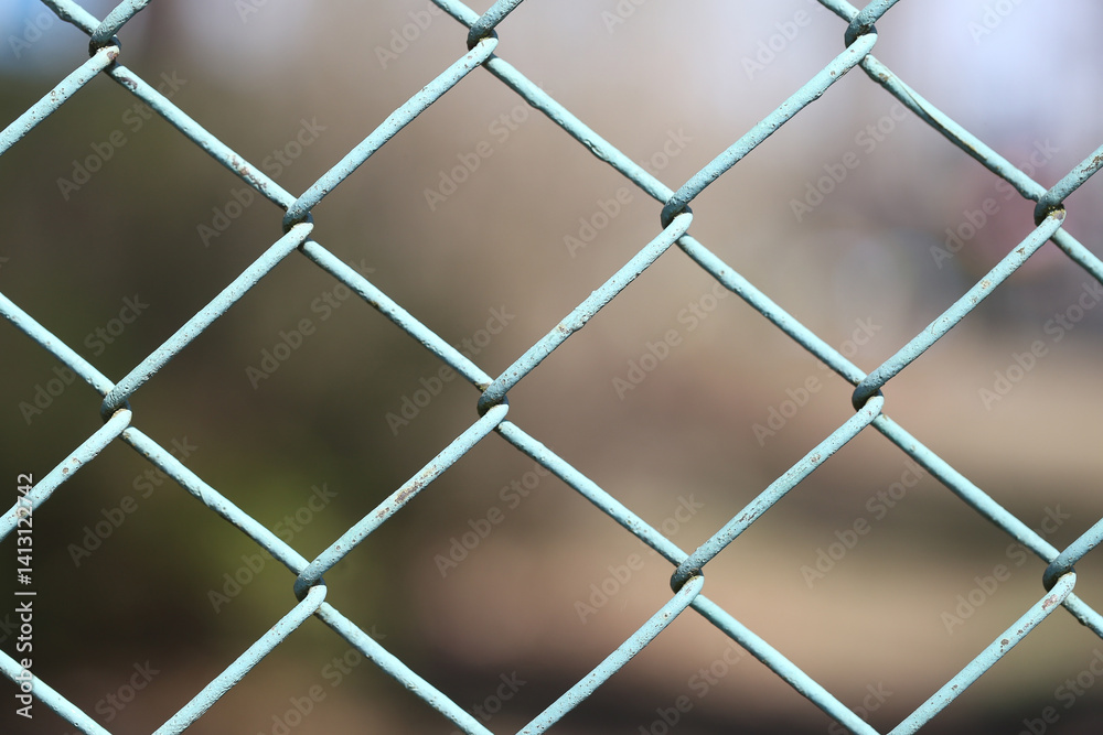 Metal fence pattern view. Grid closeup shot. Chain link mesh. Structure in focus. Blurred soft background. Blue cold steel. Industrial feel mood. Geometric metal line design.