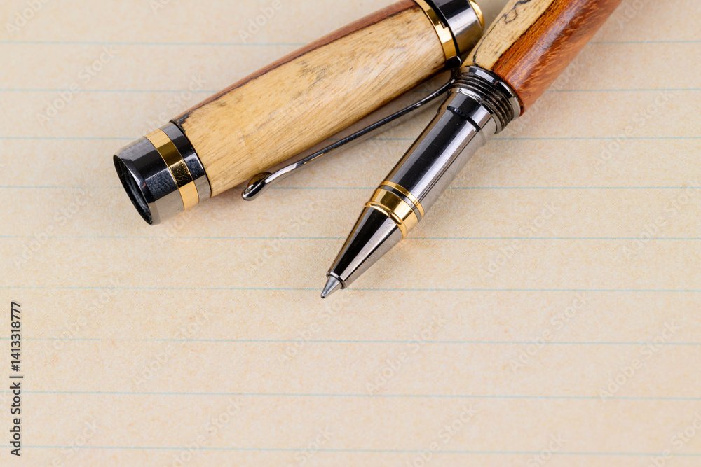 Fototapeta premium elegant wooden pens with natural grain patterns and brass and metal accents, placed on an open notebook with blank cream-colored pages, ideal for writing, office, and creative projects.