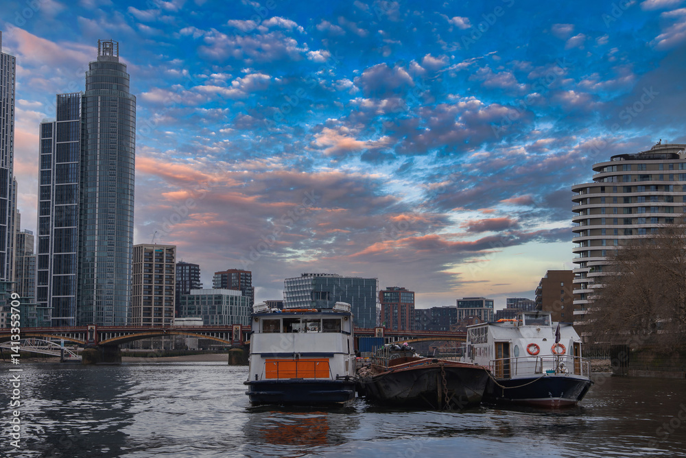 Serene view of the River Thames in London with moored boats, a glass skyscraper, a curved residential building, and a bridge under a colorful sky.