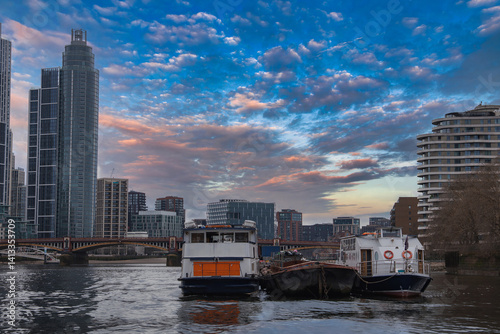 Obraz na płótnie Serene view of the River Thames in London with moored boats, a glass skyscraper, a curved residential building, and a bridge under a colorful sky. w salonie